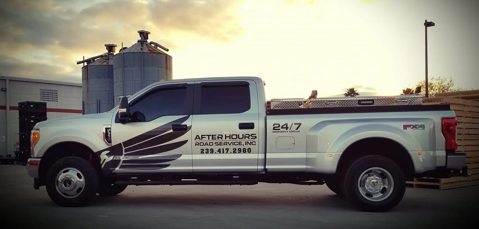 The After Hours Road Service truck: a white Ford pickup with a black wing wrap, the company name, phone number and "24/7" on the side, at sunset